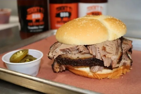A close-up of a brisket sandwich served on a bun with a side of pickled jalapenos, with bottles of Tommy T's BBQ sauce visible in the background.