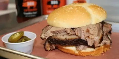 A close-up of a brisket sandwich served on a bun with a side of pickled jalapenos, with bottles of Tommy T's BBQ sauce visible in the background.