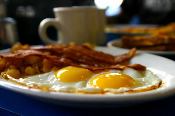 A plate of sunny-side up eggs, bacon, and hash with a coffee mug in the background.