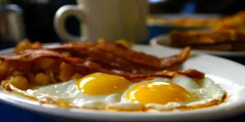 A plate of sunny-side up eggs, bacon, and hash with a coffee mug in the background.