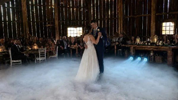A bride and groom dancing on a low-lying fog-covered dance floor in a rustic barn venue with blue uplighting.