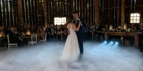 A bride and groom dancing on a low-lying fog-covered dance floor in a rustic barn venue with blue uplighting.