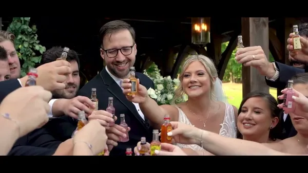 A bride and groom smiling and toasting with miniature liquor bottles surrounded by wedding guests at a reception.
