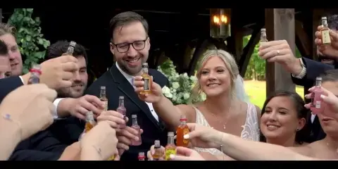 A bride and groom smiling and toasting with miniature liquor bottles surrounded by wedding guests at a reception.