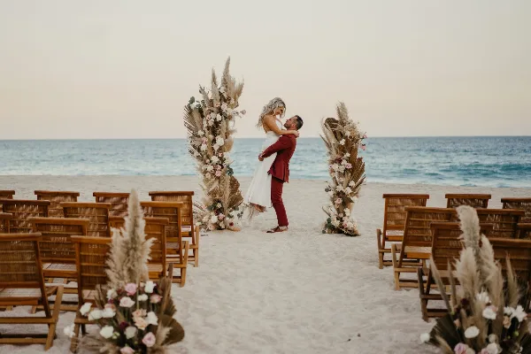 A beach wedding ceremony setup with a floral arch, wooden chairs, and a couple embracing.