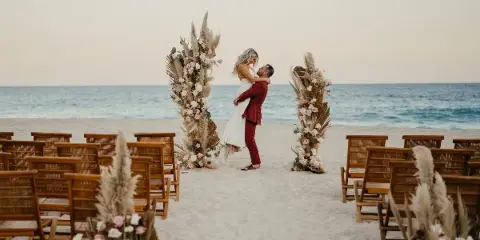 A beach wedding ceremony setup with a floral arch, wooden chairs, and a couple embracing.