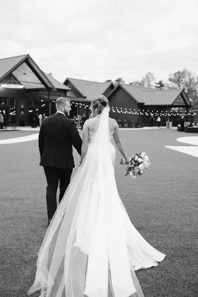 A black and white documentary-style photograph of a bride and groom walking away from the camera on a lawn at a wedding venue.