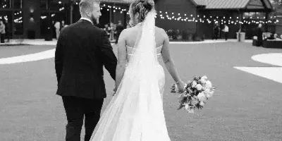 A black and white documentary-style photograph of a bride and groom walking away from the camera on a lawn at a wedding venue.