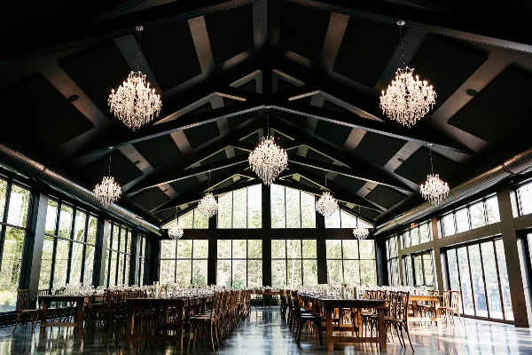 Interior view of a modern wedding reception hall with a high vaulted ceiling, crystal chandeliers, and large windows overlooking a forest.