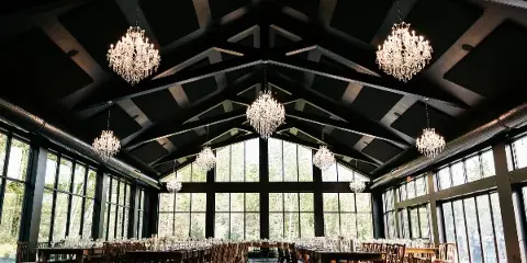 Interior view of a modern wedding reception hall with a high vaulted ceiling, crystal chandeliers, and large windows overlooking a forest.