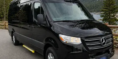Black Mercedes-Benz Sprinter van parked on a paved road with a mountain and lake in the background.