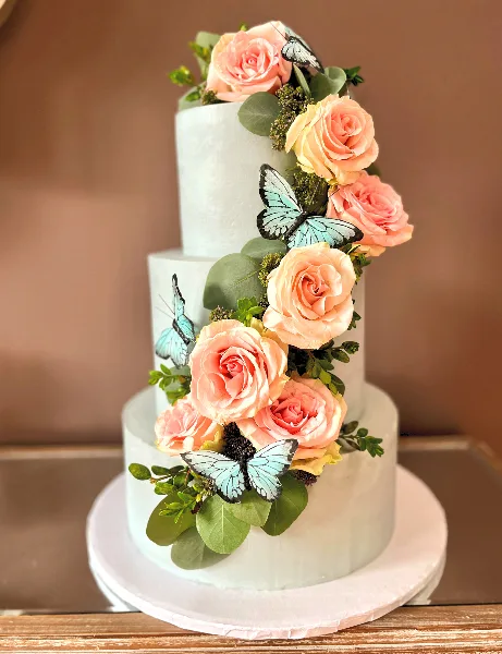 A three-tiered white wedding cake decorated with cascading pink and peach roses, greenery, and blue butterfly accents.