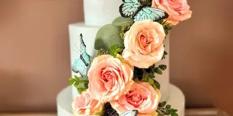 A three-tiered white wedding cake decorated with cascading pink and peach roses, greenery, and blue butterfly accents.