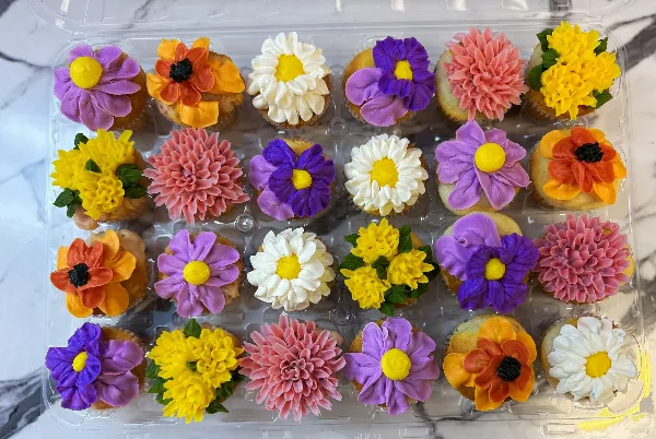 A clear plastic container holding a dozen cupcakes decorated with colorful floral frosting in purple, yellow, orange, pink, and white.