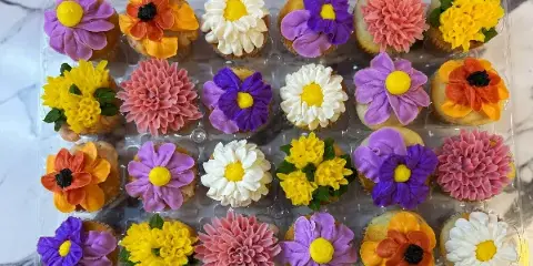 A clear plastic container holding a dozen cupcakes decorated with colorful floral frosting in purple, yellow, orange, pink, and white.