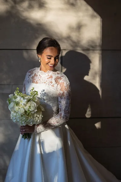 A bride in a white lace wedding dress holding a bouquet of white flowers, standing against a textured wall with dappled sunlight.