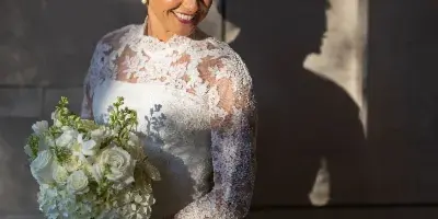 A bride in a white lace wedding dress holding a bouquet of white flowers, standing against a textured wall with dappled sunlight.