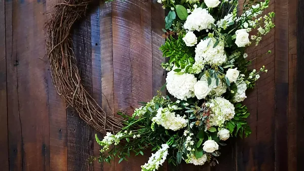 A white floral wedding arrangement with roses and hydrangeas mounted on a dark wooden panel.