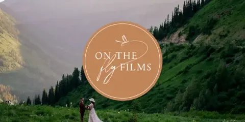 A wedding couple standing in a mountain meadow with a large 'On The Fly Films' logo overlaid in the center.