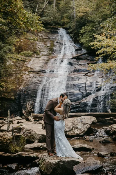 A bride and groom kissing in front of a cascading waterfall in a wooded area.