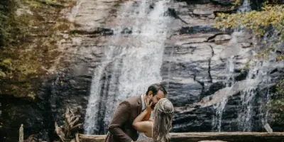 A bride and groom kissing in front of a cascading waterfall in a wooded area.