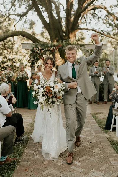 A bride and groom walking down the aisle at an outdoor wedding ceremony, smiling and waving to guests.