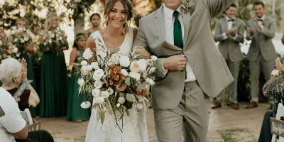 A bride and groom walking down the aisle at an outdoor wedding ceremony, smiling and waving to guests.