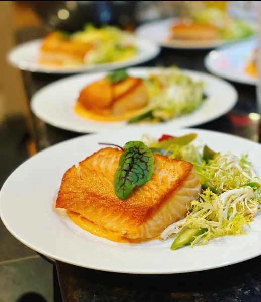 A row of plated meals featuring seared fish and salad garnishes on white plates.