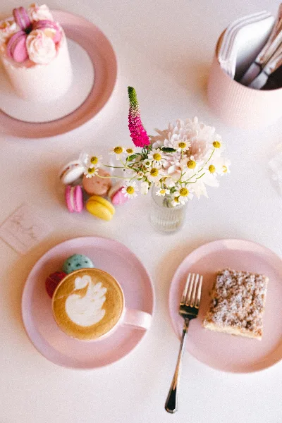 A table setting featuring a small white cake topped with macarons, a plate of colorful macarons, a latte with swan art, and a slice of cake on pink plates.