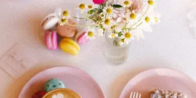 A table setting featuring a small white cake topped with macarons, a plate of colorful macarons, a latte with swan art, and a slice of cake on pink plates.