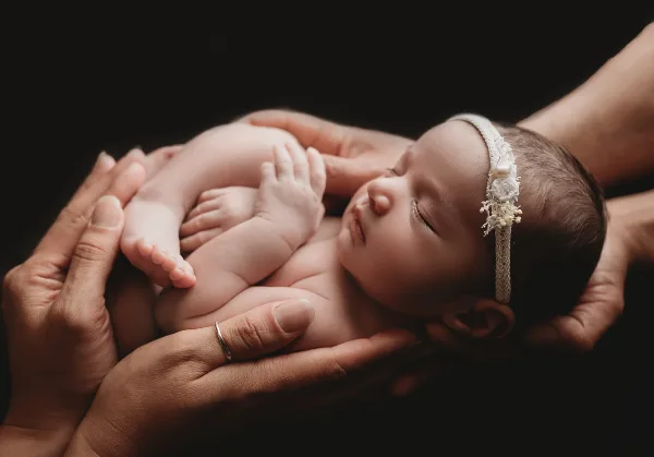 A sleeping newborn baby wearing a floral headband, cradled in the hands of parents against a dark background.