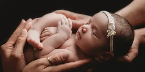 A sleeping newborn baby wearing a floral headband, cradled in the hands of parents against a dark background.