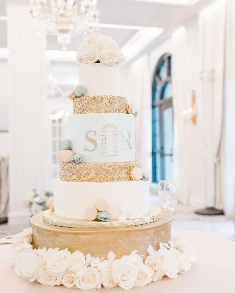 A multi-tiered white and gold wedding cake decorated with macarons and fresh flowers on a gold stand.