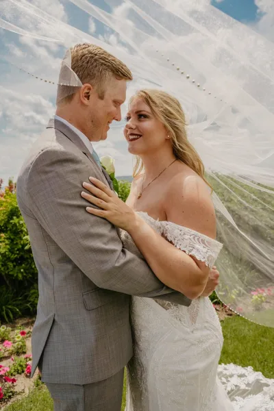 A bride and groom embracing outdoors, with the bride wearing a white lace wedding dress and the groom in a grey suit, set against a backdrop of greenery and a blue sky.