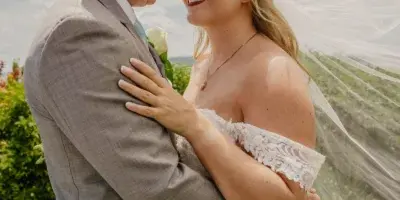 A bride and groom embracing outdoors, with the bride wearing a white lace wedding dress and the groom in a grey suit, set against a backdrop of greenery and a blue sky.