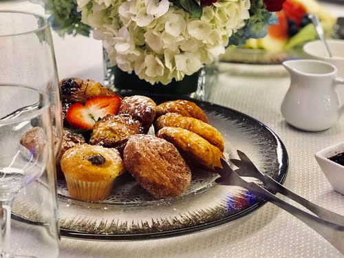 A close-up of a plated dessert assortment featuring muffins and pastries on a silver charger, set on a table with a white floral centerpiece.