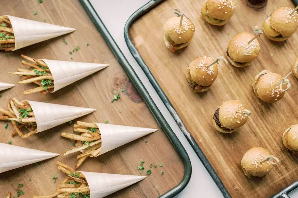 A top-down view of catering food including mini sliders on a wooden tray and french fries in paper cones on a wooden board.