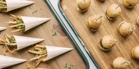 A top-down view of catering food including mini sliders on a wooden tray and french fries in paper cones on a wooden board.