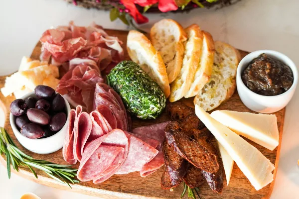 A wooden board filled with assorted cured meats, cheeses, bread, olives, and a green herb ball.