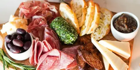 A wooden board filled with assorted cured meats, cheeses, bread, olives, and a green herb ball.