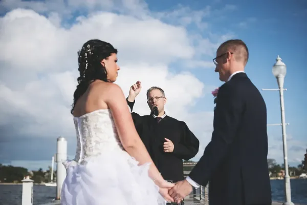 A wedding officiant in a black robe stands between a bride and groom, leading a ceremony outdoors near a body of water.