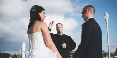 A wedding officiant in a black robe stands between a bride and groom, leading a ceremony outdoors near a body of water.