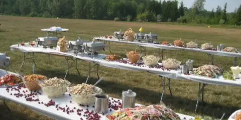 An outdoor catering buffet featuring long tables with chafing dishes, salads, and various food items set up in a grassy field.