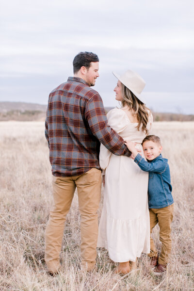 A family of three standing in a dry grassy field, with the father in a plaid shirt, mother in a white dress and hat, and a young boy in a denim jacket.