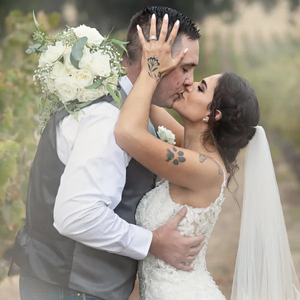 A bride and groom sharing a kiss in an outdoor setting, with the bride holding a white rose bouquet.