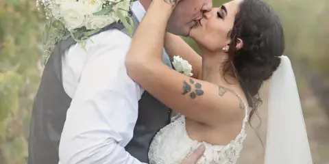 A bride and groom sharing a kiss in an outdoor setting, with the bride holding a white rose bouquet.