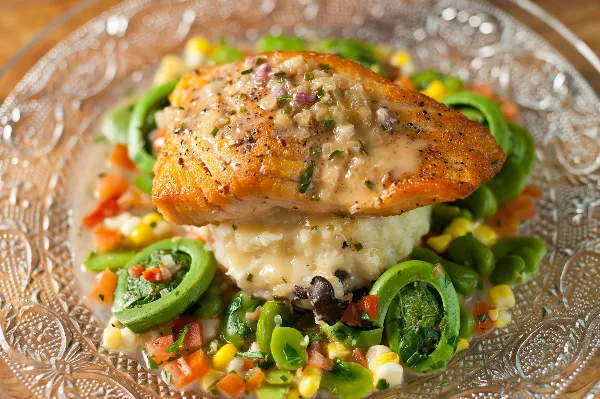 A plated salmon dish served over mashed potatoes with a side of seasonal vegetables and greens.