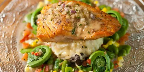 A plated salmon dish served over mashed potatoes with a side of seasonal vegetables and greens.