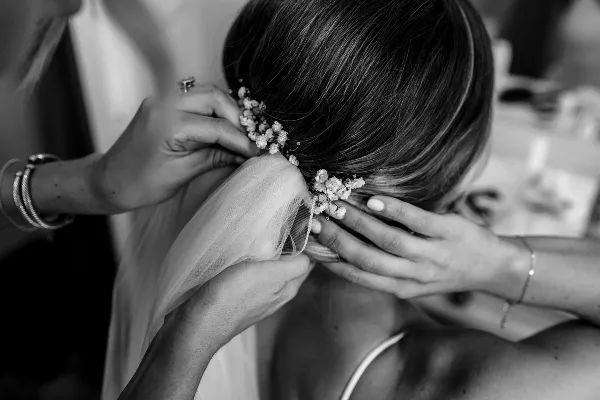 A close-up black and white photo of a hair stylist's hands arranging a bride's hair and attaching a floral accessory.