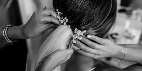 A close-up black and white photo of a hair stylist's hands arranging a bride's hair and attaching a floral accessory.